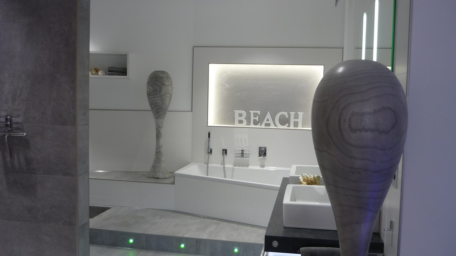 Weißes Bad mit Holzvasen und beleuchteter Nische mit BEACH-Schriftzug