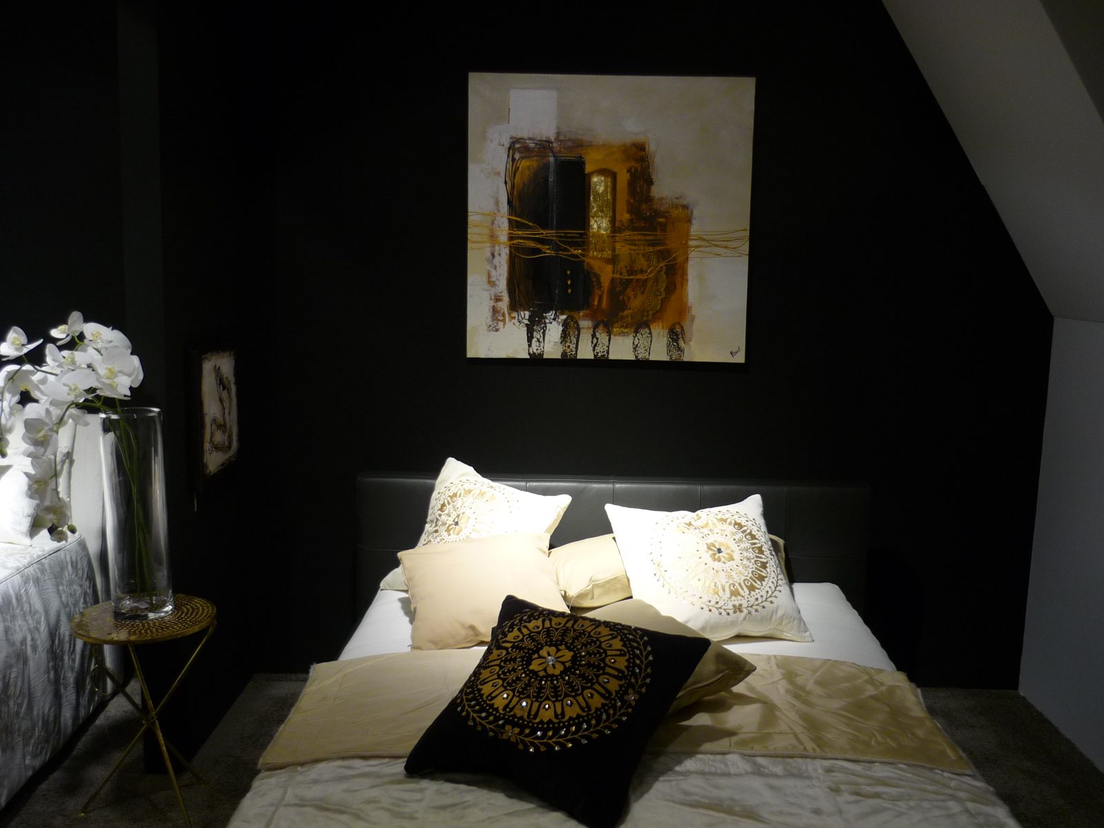 Bett vor dunkler Wand mit Gold-Ornamentkissen, Gemälde und weißen Orchideen